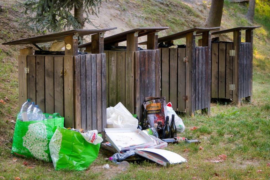 Four wooden outdoor rubbish bins with sloped, weathered dark brown lids are positioned in a row on a grassy area, surrounded by trees and a slight hillside. The bins are made of vertical wooden planks